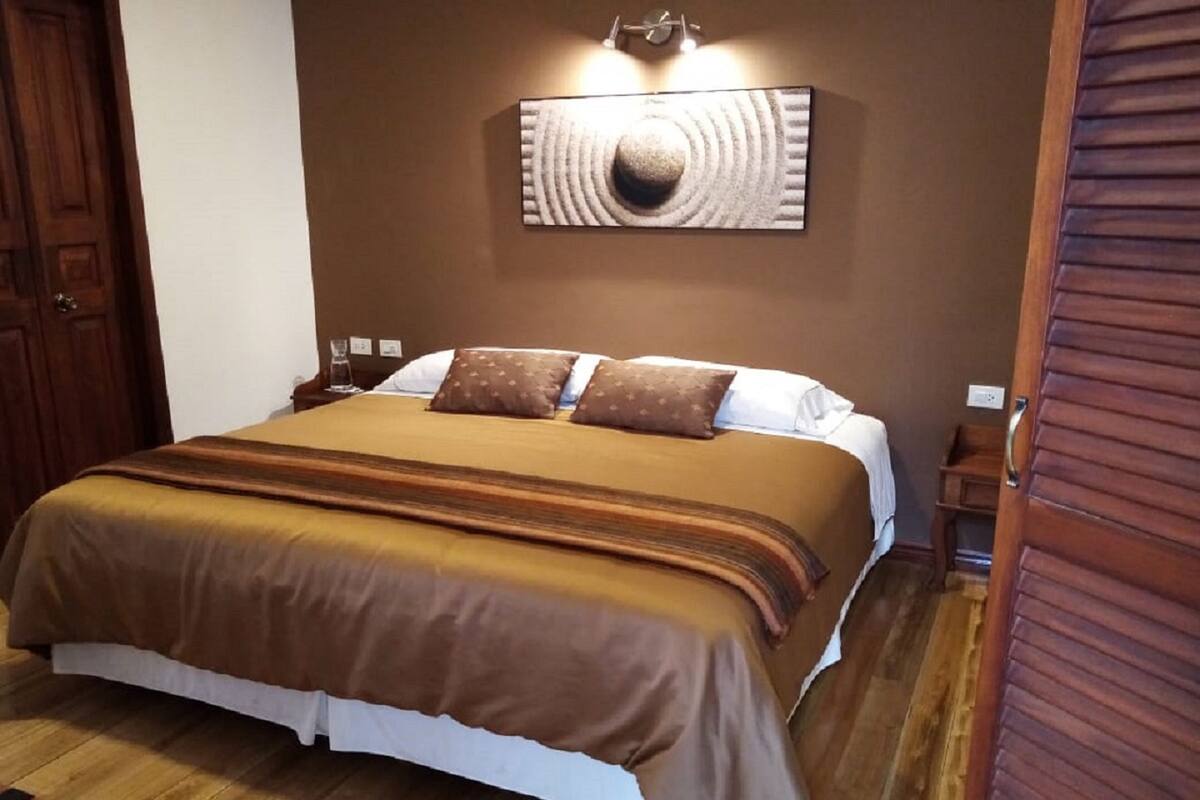 Ein ordentlich bezogenes Bett mit brauner Tagesdecke und Kissen, Holzboden und eine wandmontierte Lampe.