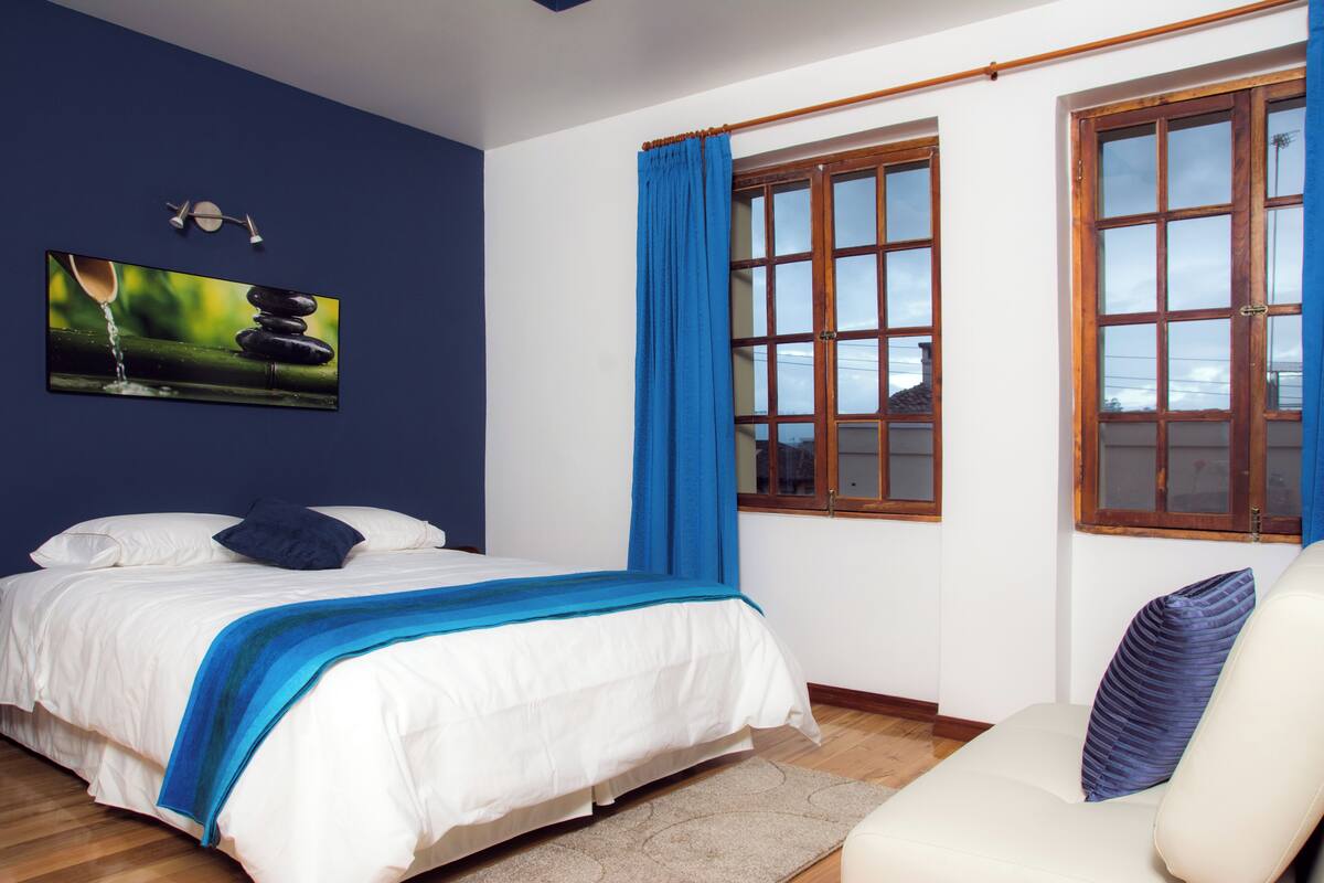 Ein Schlafzimmer mit Bett, Stuhl, einem Fenster mit Vorhängen und einem Gemälde an der Wand.