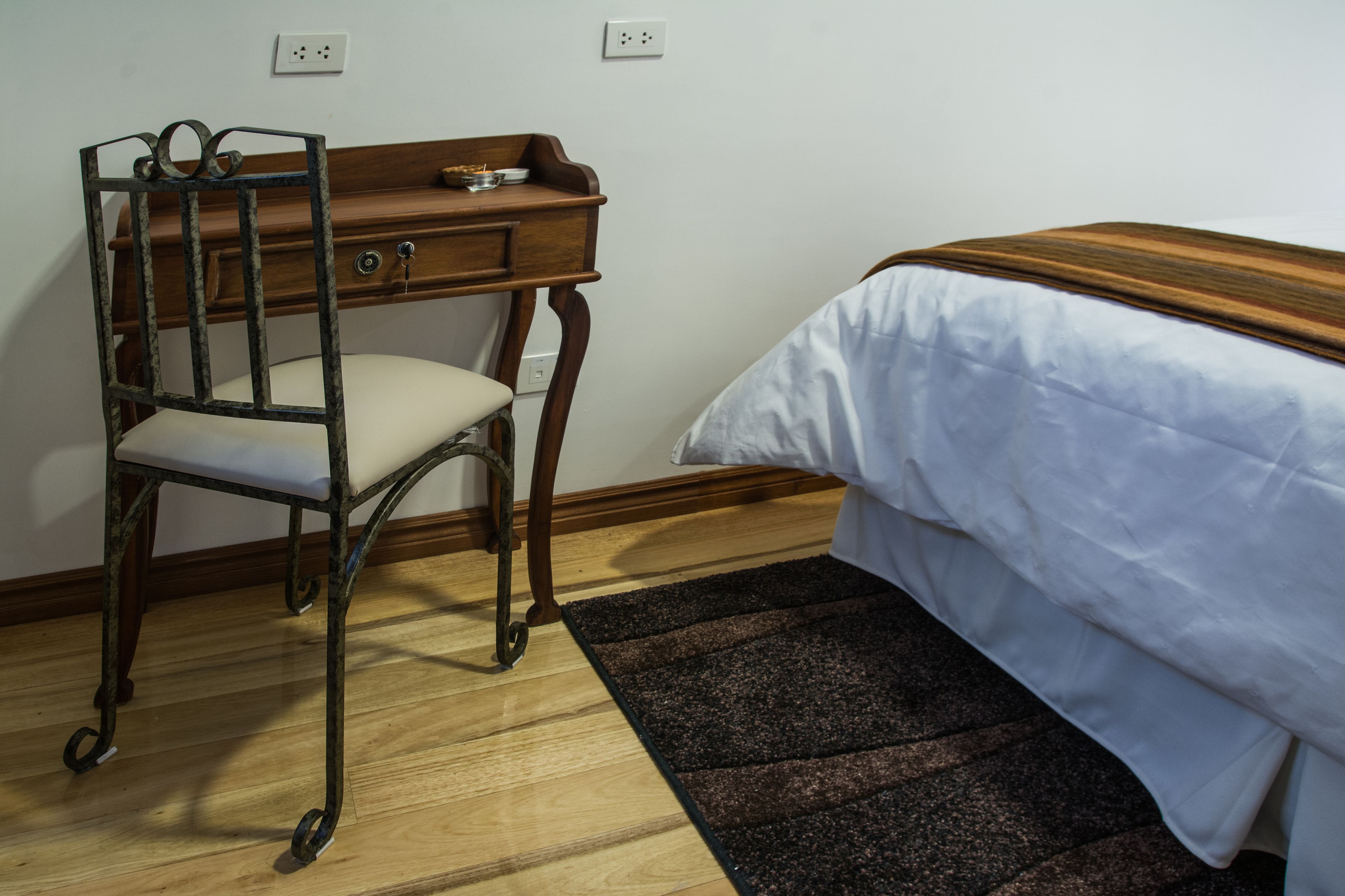 Ein Zimmer mit einem Holztisch, einem Sessel, einem Bett mit weißer Bettdecke und einem dunkelbraunen Teppich.