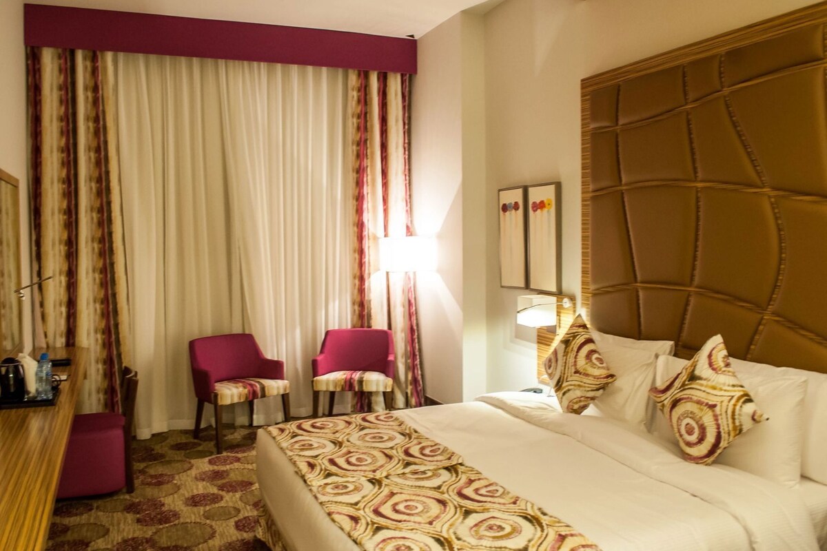A hotel room with a bed, two chairs, a desk, and a large window with curtains.