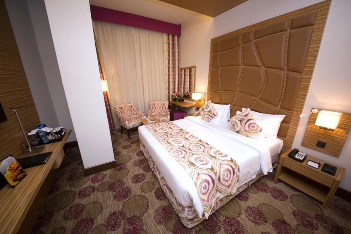 A hotel room with a large bed, a wooden headboard, a bedside table with a lamp, a chair, and a desk with a mirror.