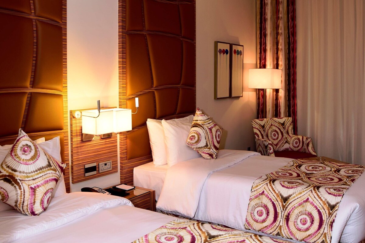A hotel room with two beds, a wooden headboard, a bedside table with a lamp, and a large window with curtains.