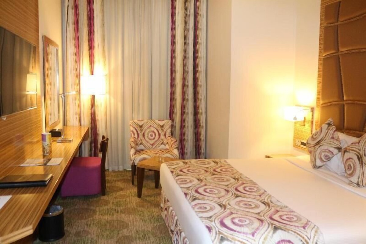 A hotel room with a bed, a desk, a chair, a small stool, and a mirror.