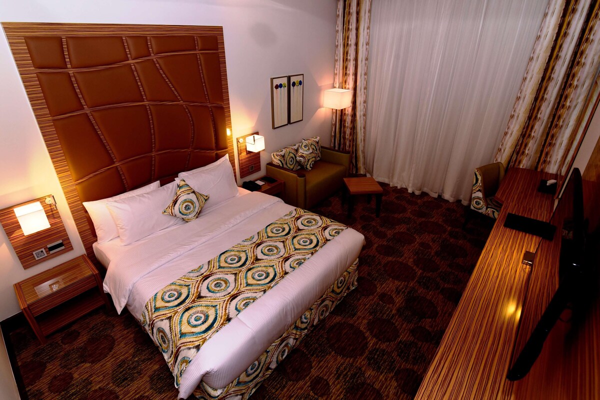 A hotel room with a large bed, a bedside table, a nightstand, a small table, a chair, a desk, and a lamp.