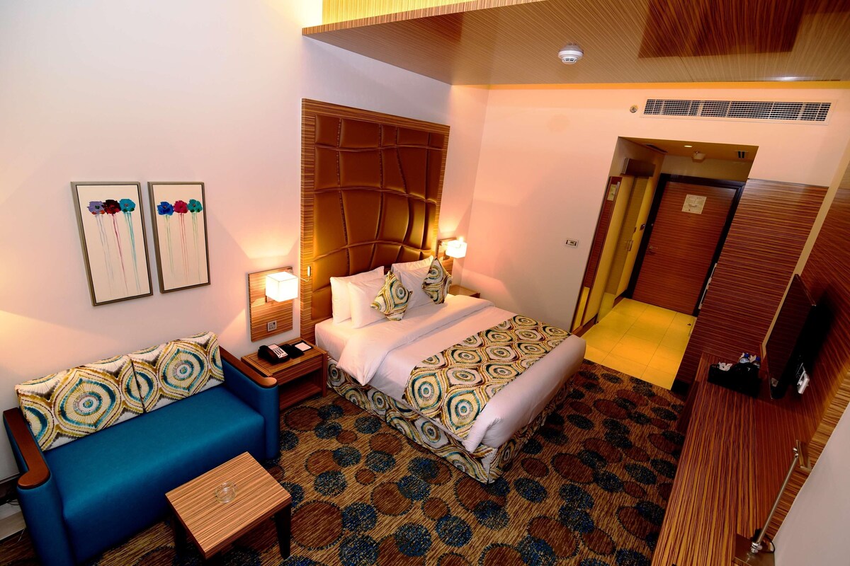 A hotel room with a bed, a blue sofa, a wooden nightstand, and a patterned carpet.