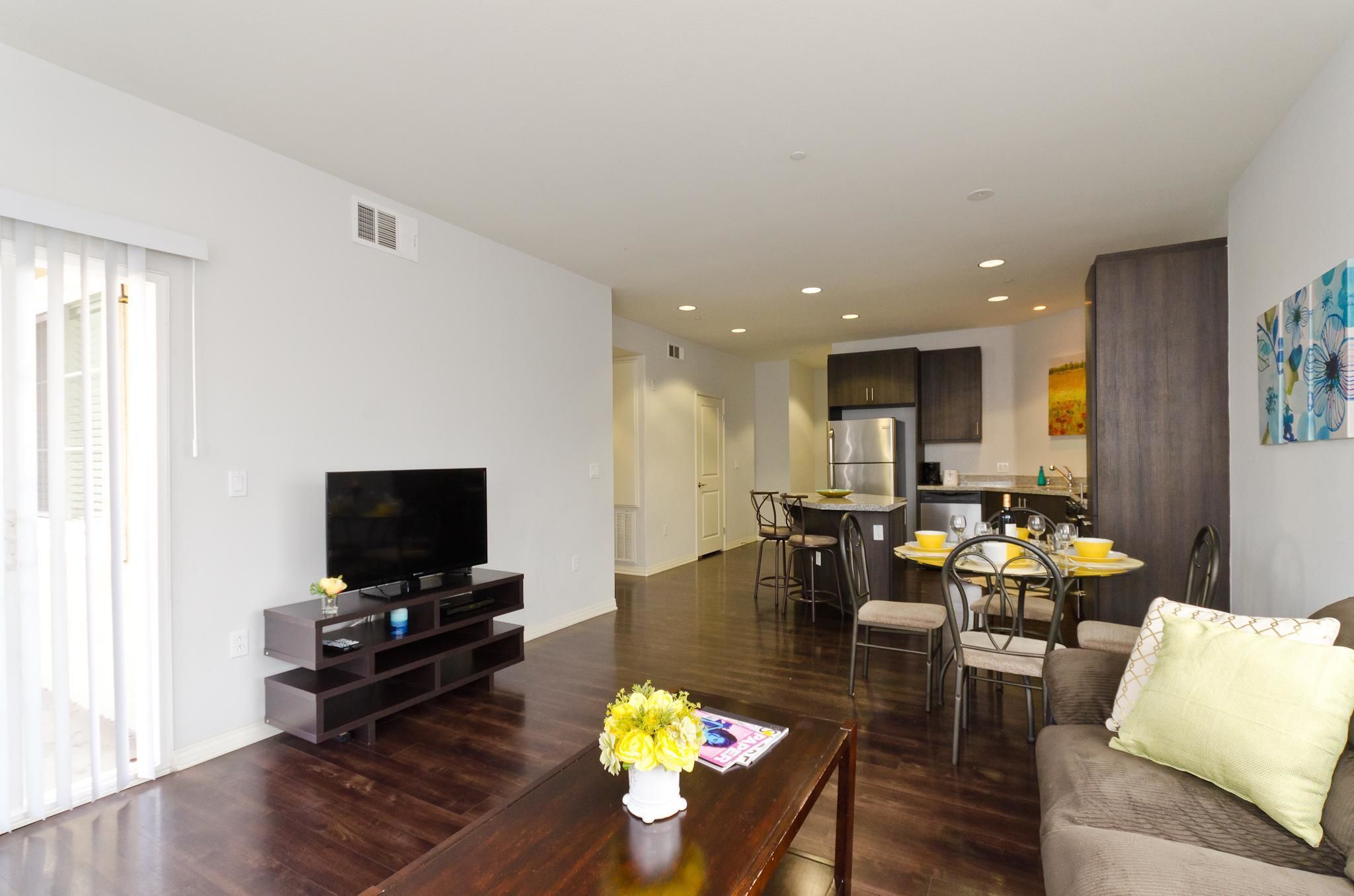 A modern living room with a flat-screen TV, a coffee table with a vase, a sofa, and a dining area with chairs and a table.