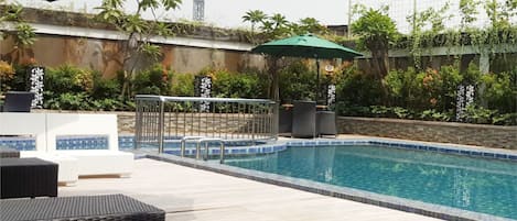 Zona de piscina con sillas de descanso y sombrillas, rodeada de vegetación y un muro decorativo.