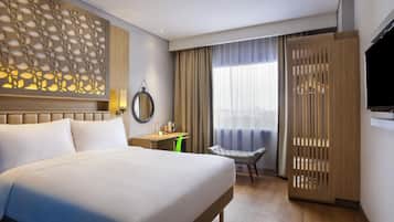Habitación de hotel con una cama grande, un escritorio, una silla, un televisor y vistas de la ciudad a través de la ventana.