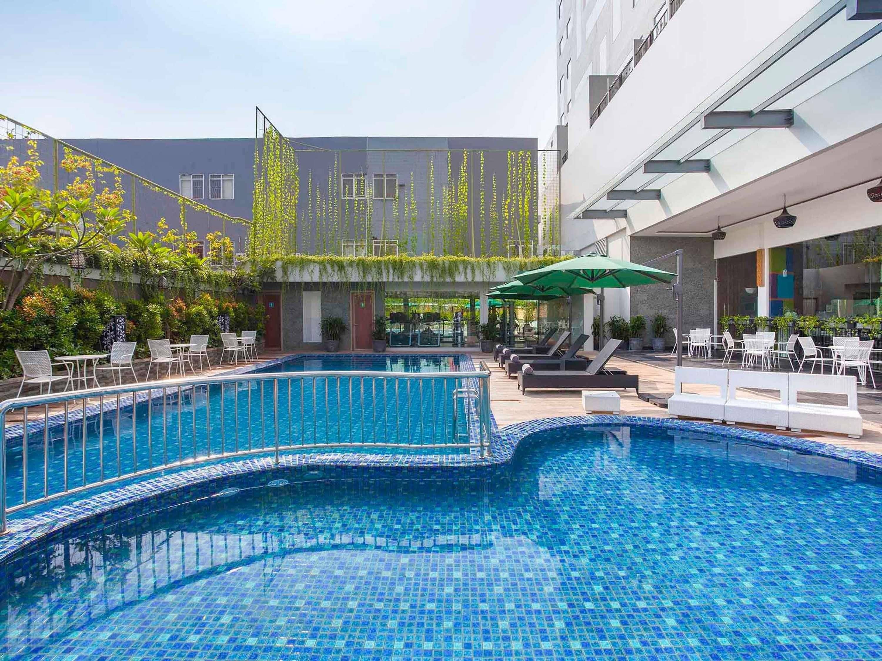 A swimming pool with lounge chairs and umbrellas, surrounded by greenery and modern buildings.