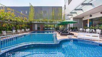 Una piscina con sillas de descanso y sombrillas, rodeada de vegetación y edificios modernos.