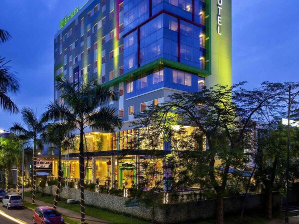 A multi-story hotel with a modern design, featuring a prominent vertical sign, a well-lit entrance, and a landscaped area with palm trees.