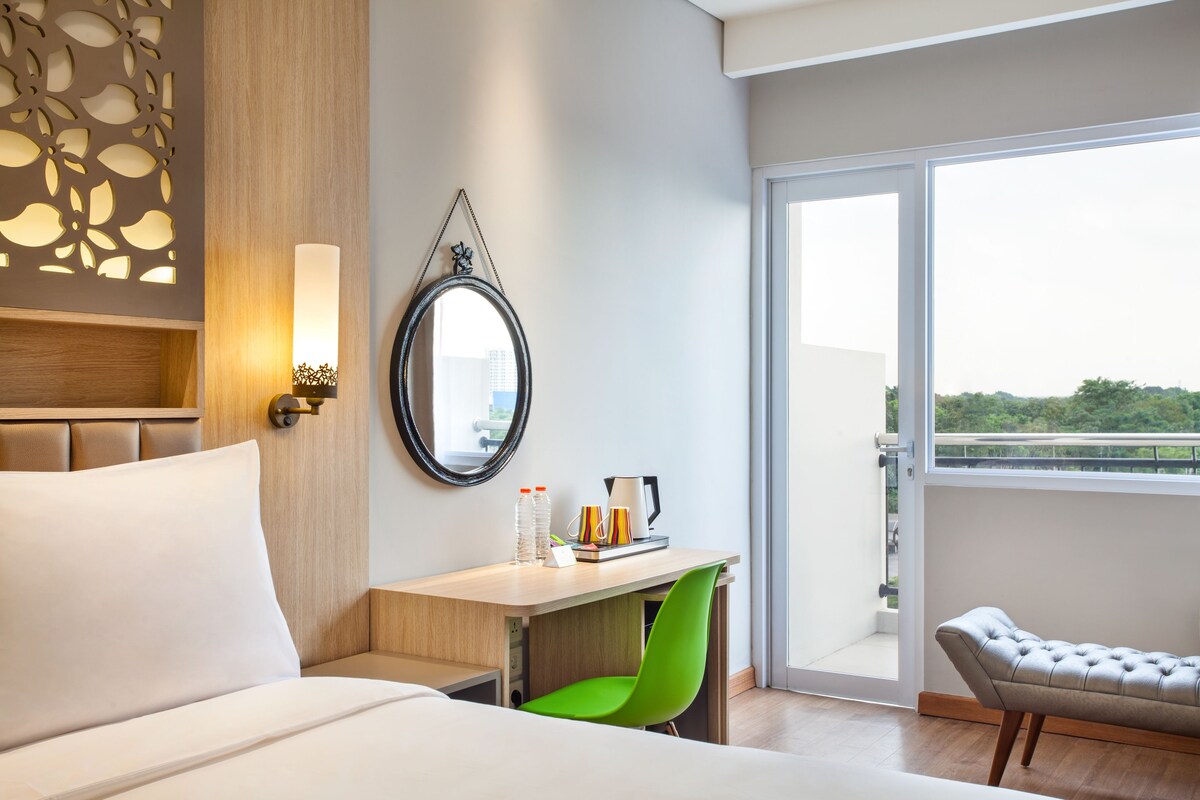 A hotel room with a bed, a desk with a chair, a small table with a kettle and cups, a mirror, and a balcony with a view.