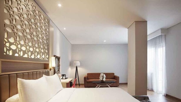Quarto de hotel com uma cama grande, um sofá, uma mesinha e um painel decorativo na parede.