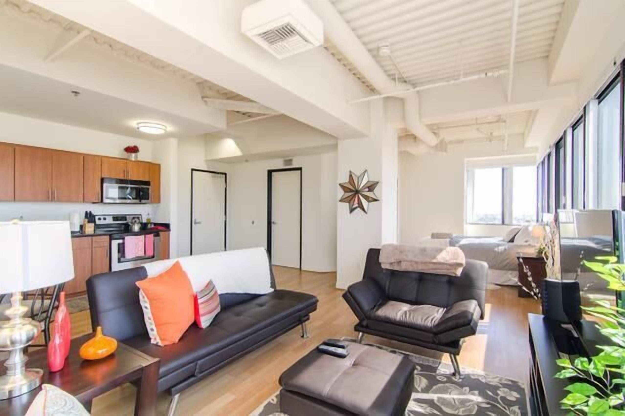 A modern living room with a black sofa, a small table, and a television. There is a kitchen area with wooden cabinets and a microwave. The room has a large window providing natural light.