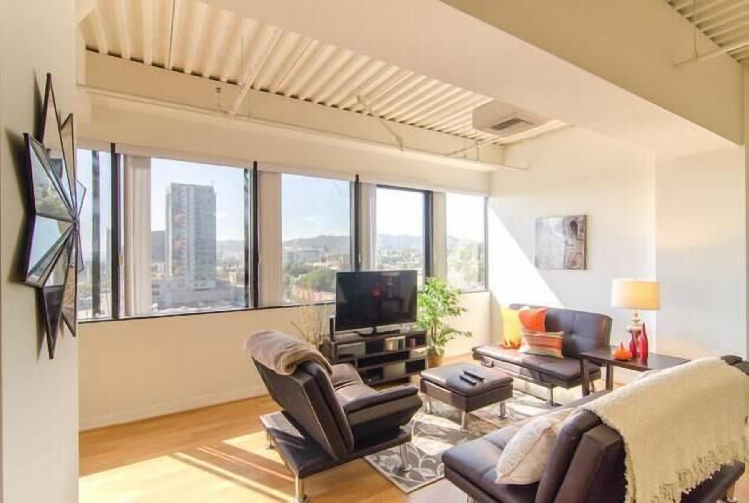 A modern living room with a flat-screen TV, comfortable seating, and large windows offering a city view.