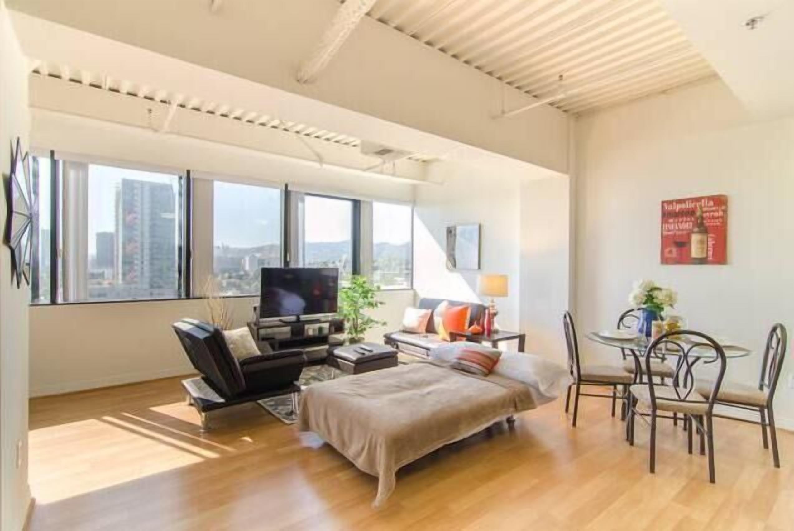 A modern hotel room with a large bed, a TV, a dining area, and a city view.