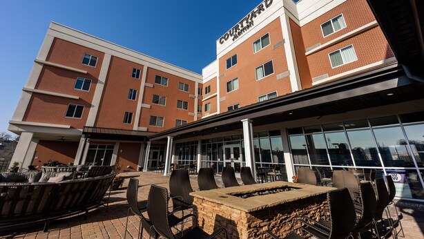 A multi-story hotel with a prominent sign reading "Courtyard." It features an outdoor seating area with chairs and a stone table, large windows, and a clear blue sky. - Courtyard by Marriott Rome (Rome)