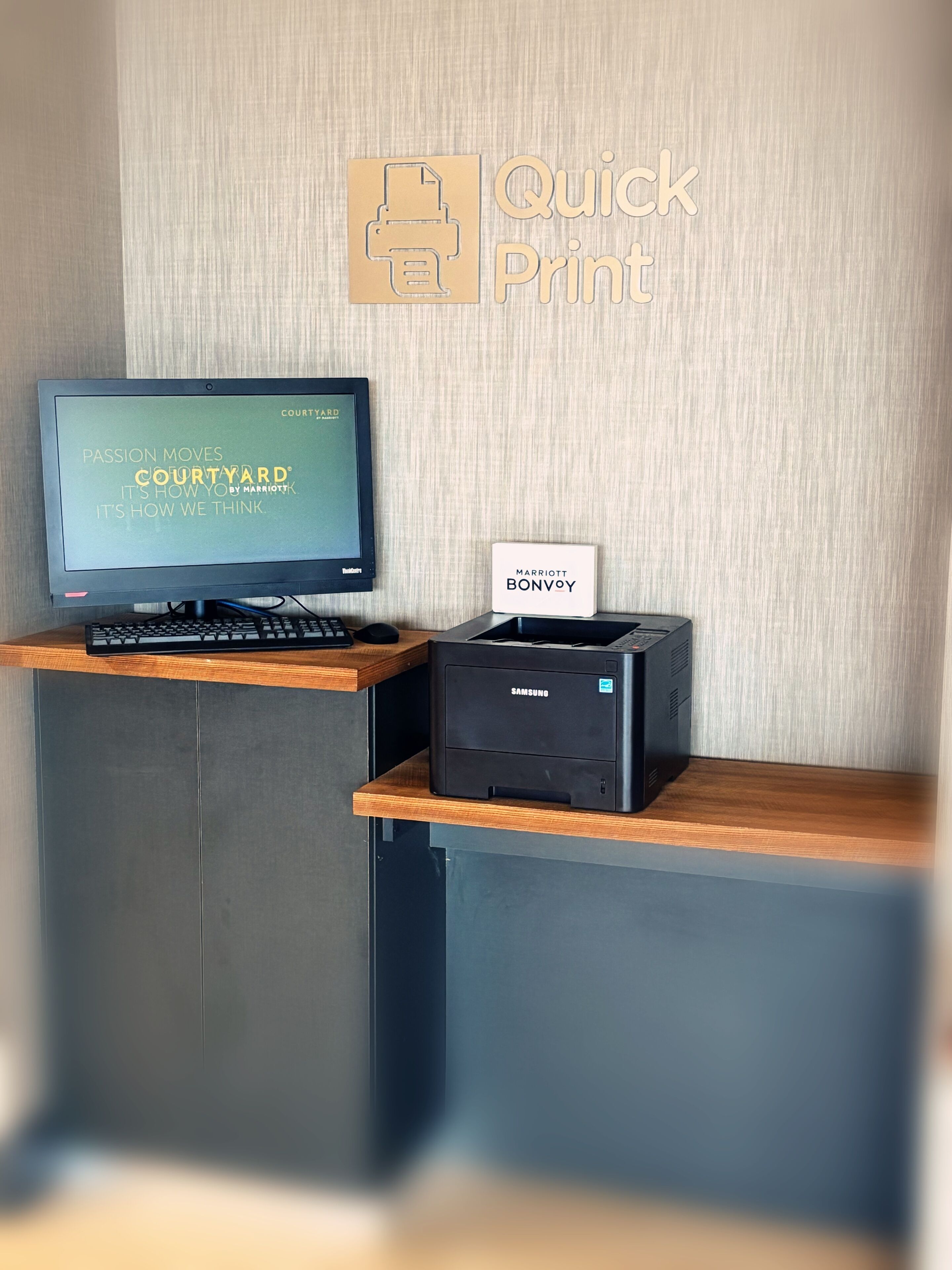 Un mostrador de recepción de hotel con un monitor que muestra la marca 'COURTYARD', una impresora Samsung y un cartel que dice 'MARRIOTT BONVOY'.