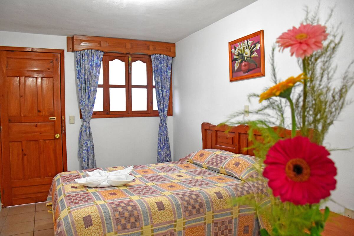 A bedroom with a wooden door, a window with curtains, a bed with a patterned bedspread, and a painting on the wall.
