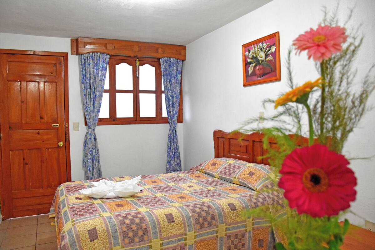 A bedroom with a wooden door, a window with curtains, a bed with a patterned bedspread, and a painting on the wall.