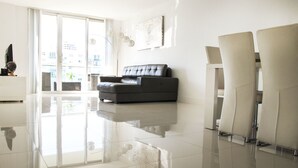 A modern living room with a black leather sofa, a white coffee table, and a large window with a view of buildings. - Sunny Isles Suites (Sunny Isles Beach)
