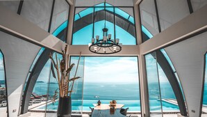 A modern interior with a large window overlooking a pool and the sea, a chandelier, and a dining table with chairs. - Sky Dream Villa (Koh Samui)