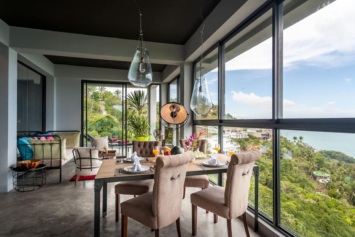 Un espace repas avec une grande table dressée pour quatre personnes, entouré de grandes fenêtres offrant une vue sur un paysage luxuriant et une étendue d’eau.
