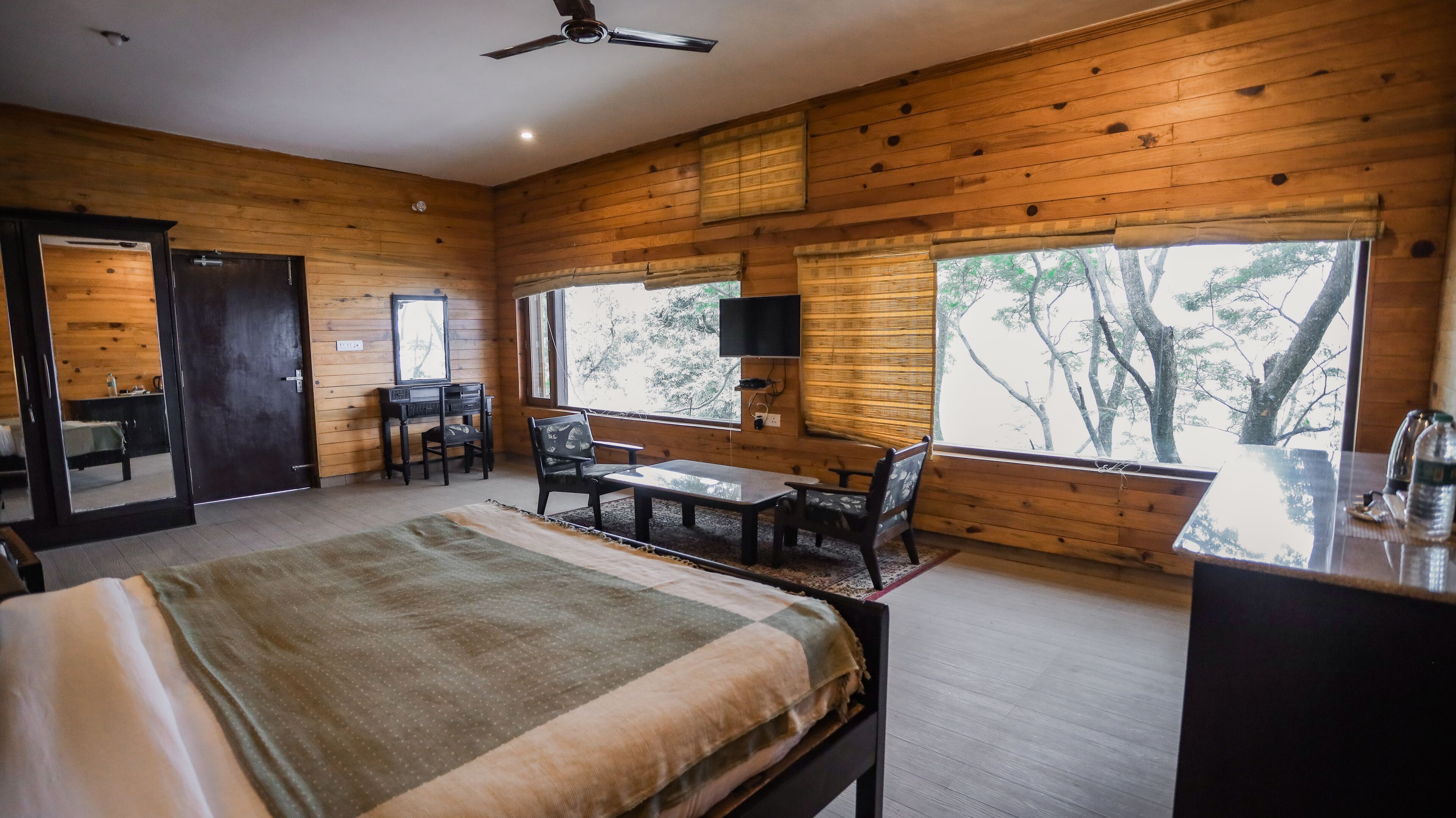 A wooden cabin room with a bed, a desk, a chair, and a large window offering a view of trees.