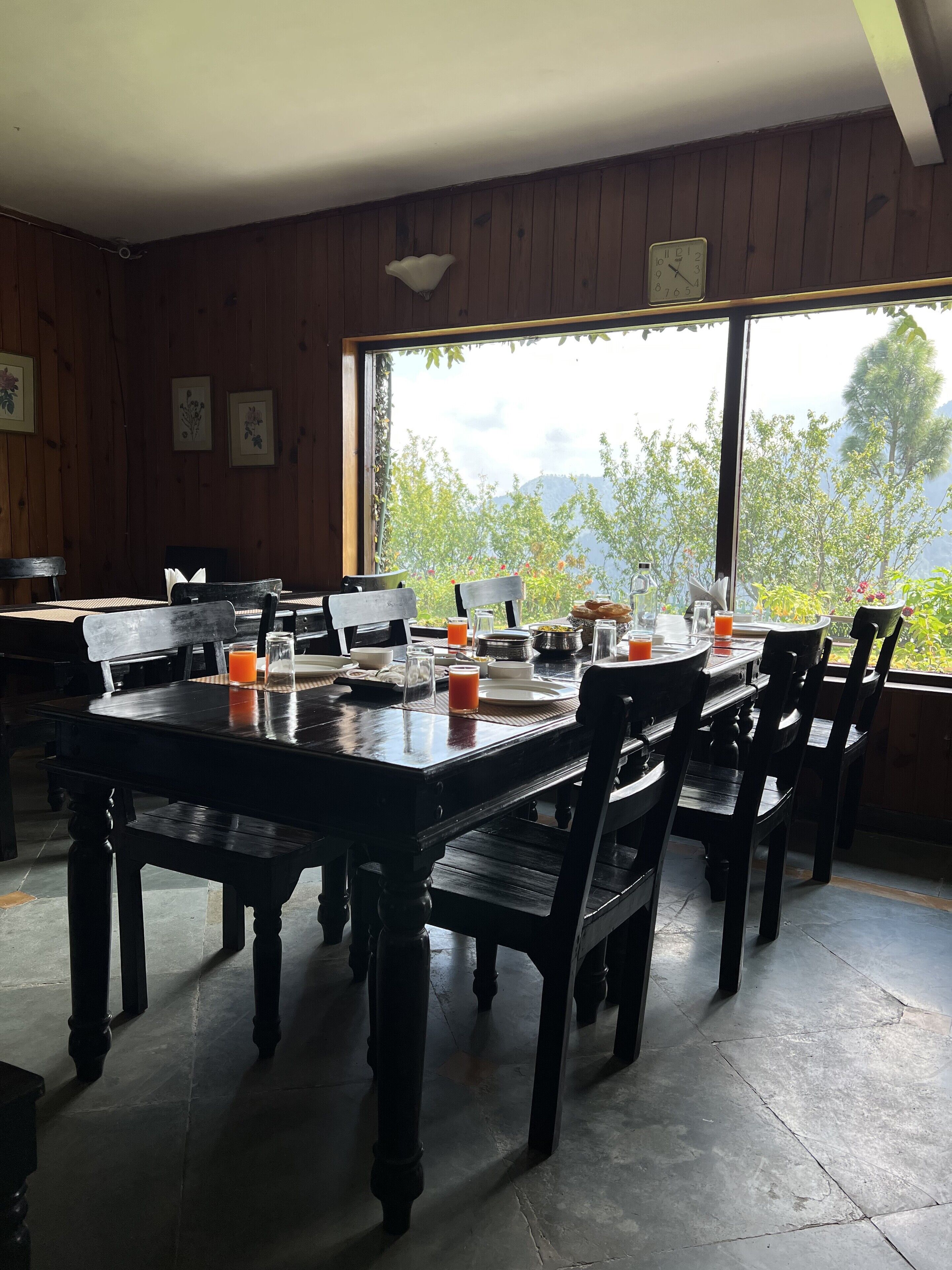 Un espace repas avec des tables et des chaises en bois, une vue sur la verdure et les montagnes grâce à de grandes fenêtres, et une horloge au mur.