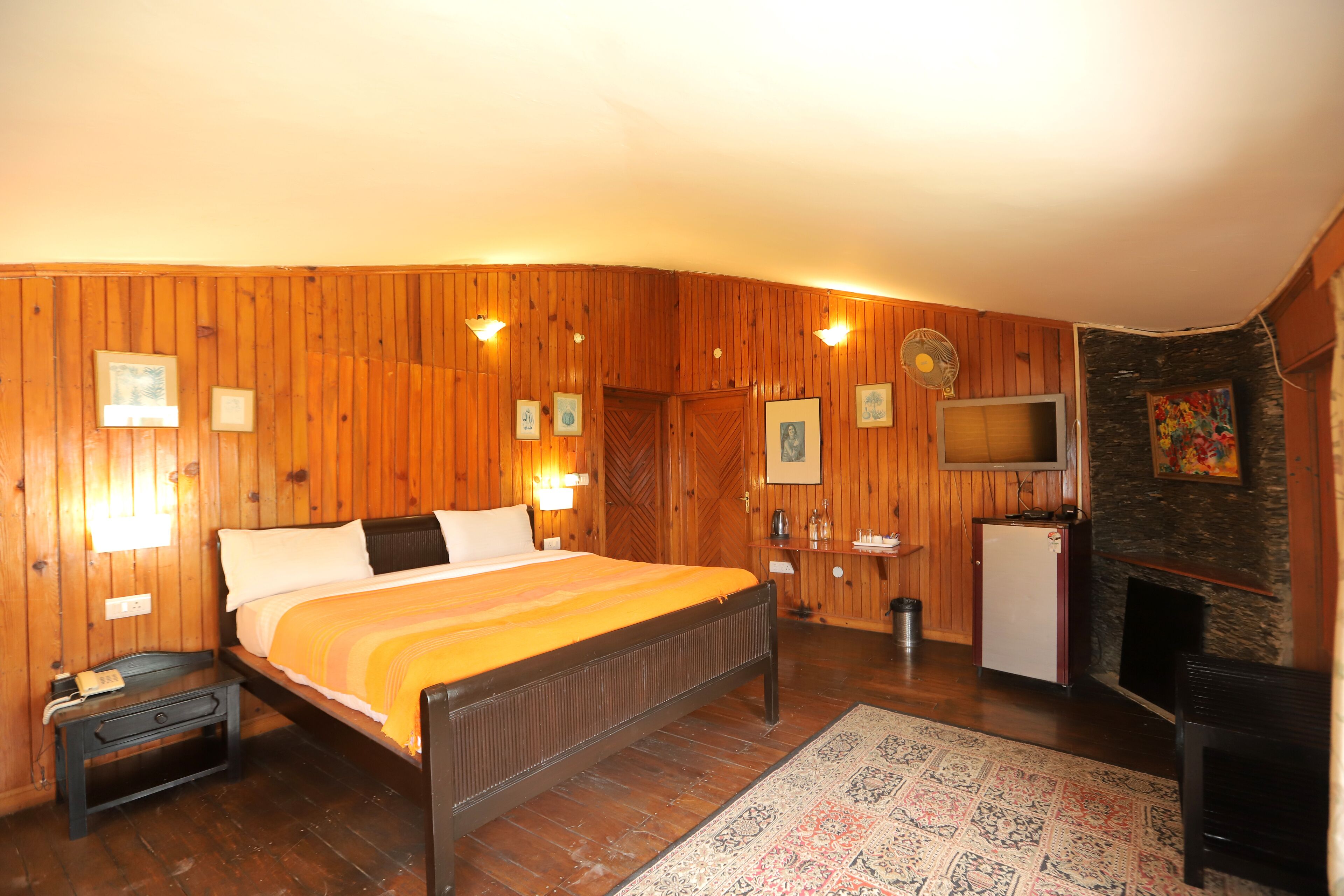A wooden-paneled bedroom with a bed, bedside table, lamp, TV, and fireplace.