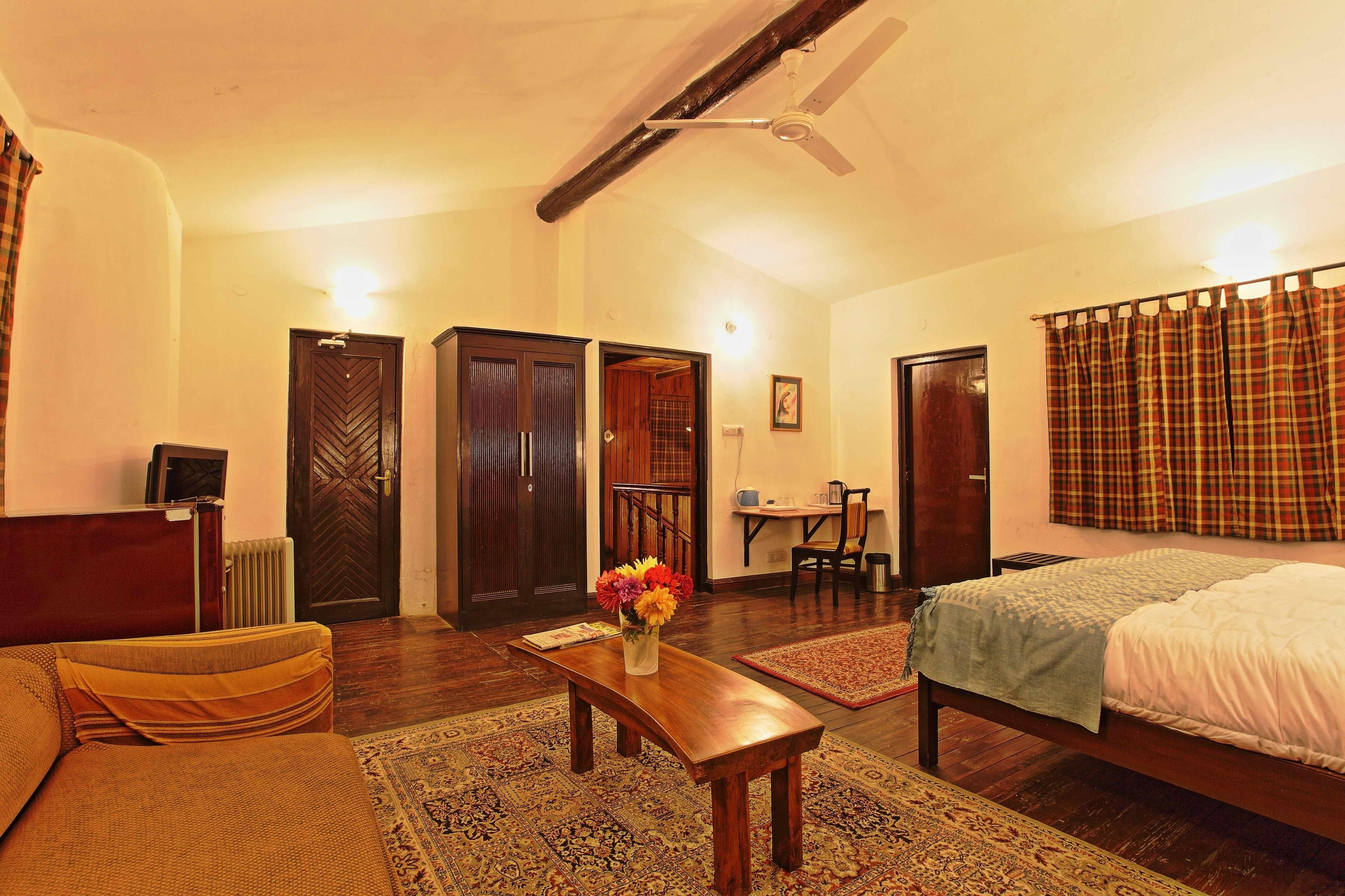 A room with a bed, a wooden bench, a small table with a vase of flowers, a radiator, a desk, and a chair.