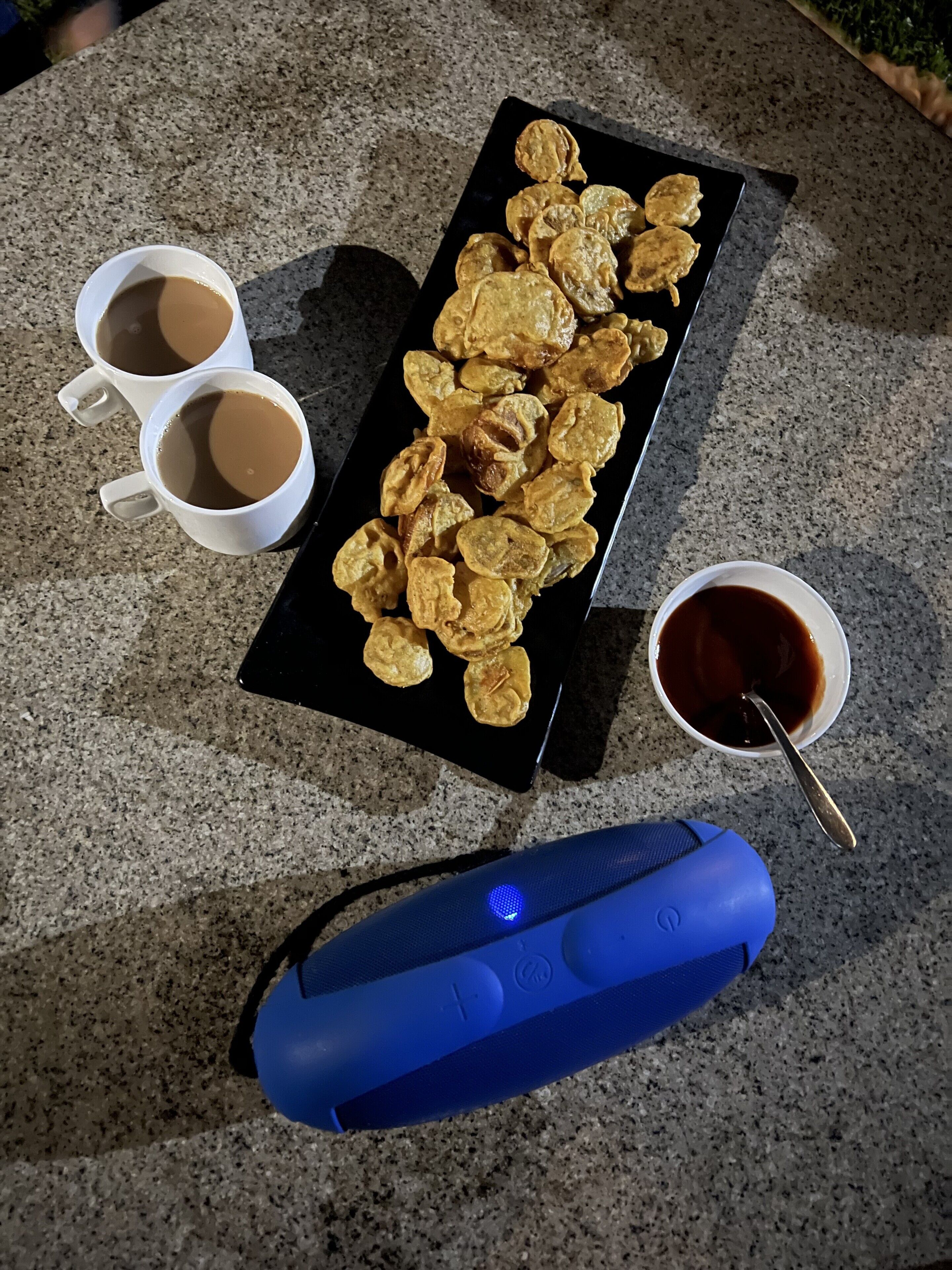 A table with a black tray holding fried snacks, two white cups of coffee, a small bowl of sauce, and a blue object.
