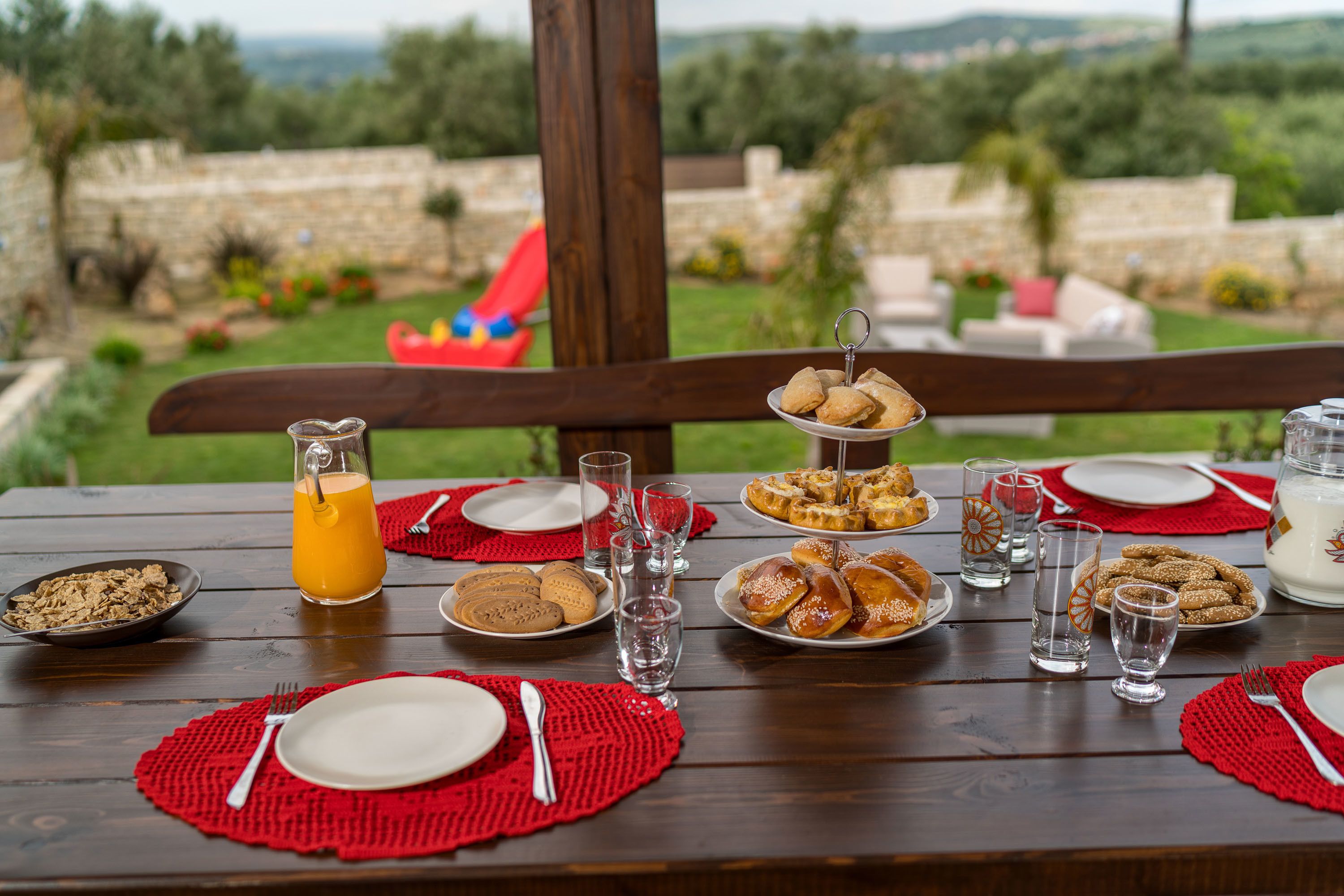 Una mesa de madera dispuesta para el desayuno, con pasteles y una jarra de jugo de naranja, con vista a un jardín con muro de piedra y vegetación.