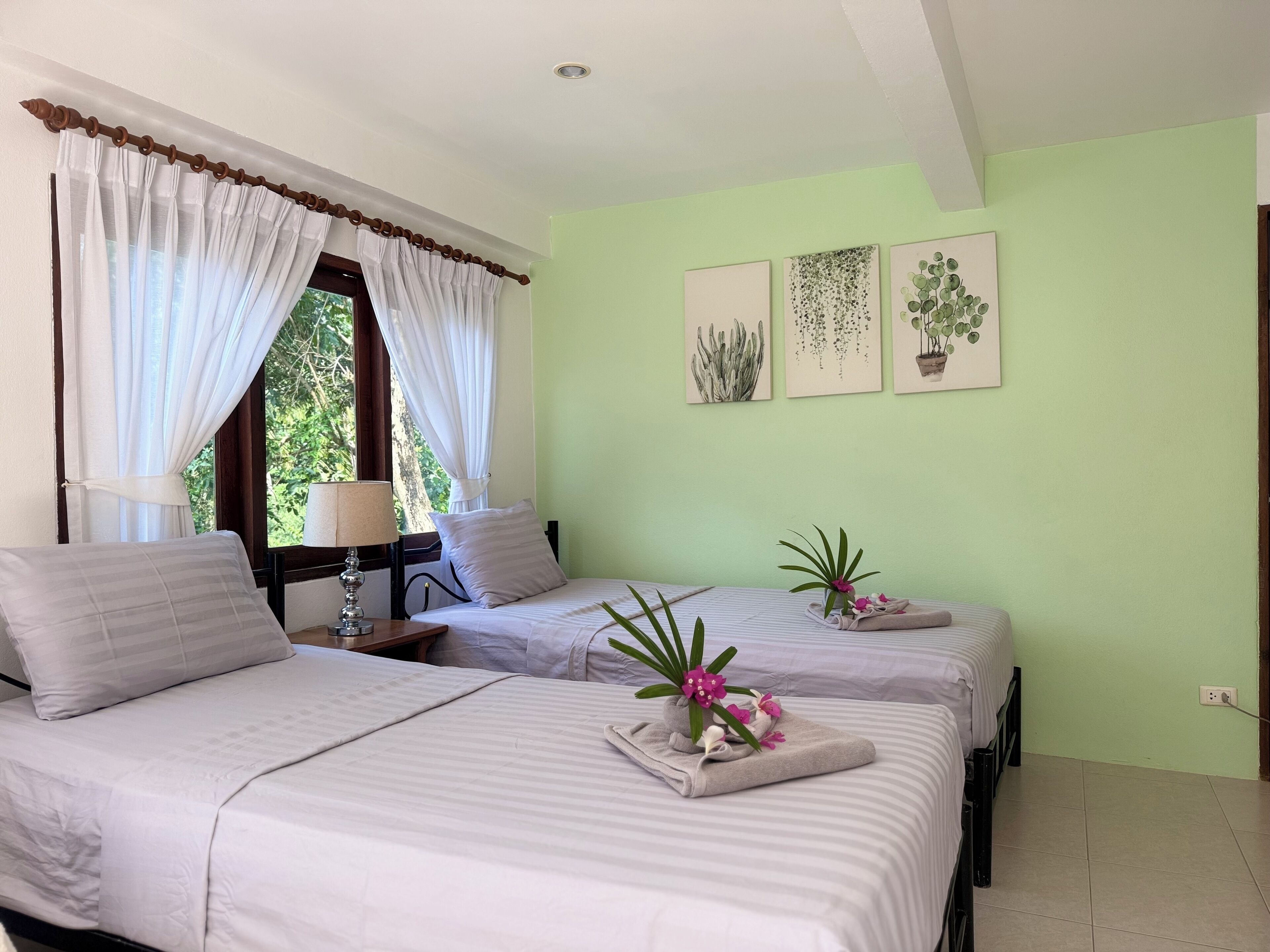 Habitación de hotel con dos camas, una ventana con cortinas, una mesita de noche con lámpara y una obra de arte en la pared que representa plantas.