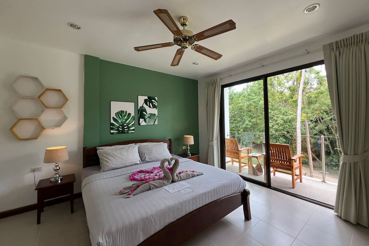 Un dormitorio con cama, ventilador de techo, mesita de noche, lámpara, ventana con vistas a zonas verdes y balcón con sillas.