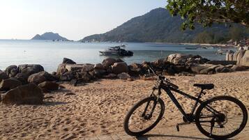 Una bicicleta estacionada en una playa arenosa con rocas grandes, un litoral rocoso y una vista al mar y montañas al fondo.