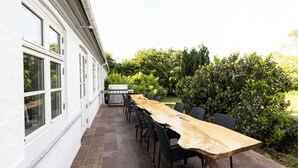 An outdoor patio with a long wooden table and chairs, a barbecue grill, and a white building with large windows.