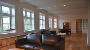 A spacious living room with a leather sofa, wooden coffee table, and a dining area with a table and chairs. Large windows provide natural light. - Lehnsgaard Ranch (Gudhjem)