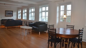 A spacious living area with a wooden floor, a round dining table with chairs, a sofa, and a small white table. - Lehnsgaard Ranch (Gudhjem)