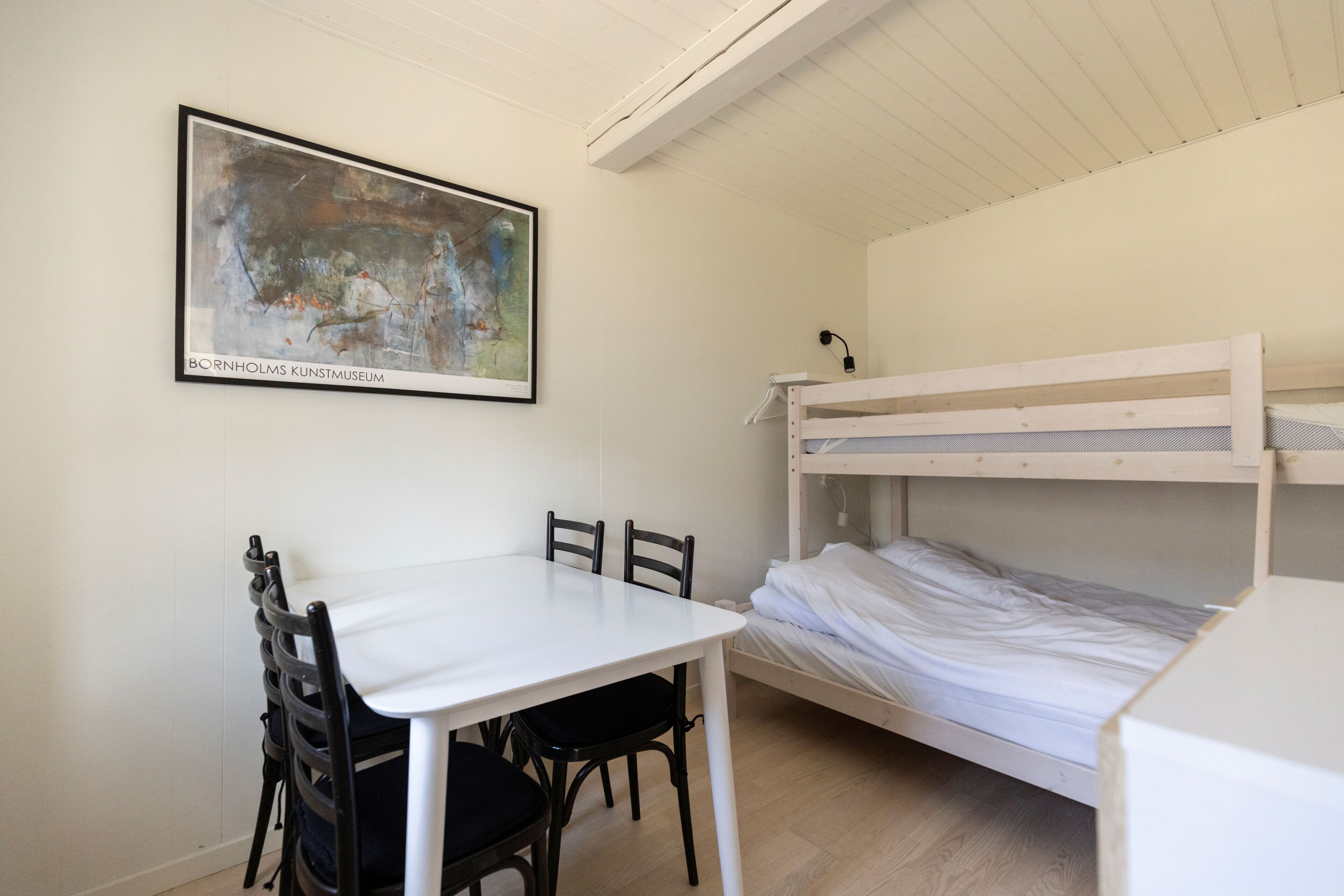 A bunk bed with a white mattress, a white table with chairs, and a framed abstract painting on the wall.