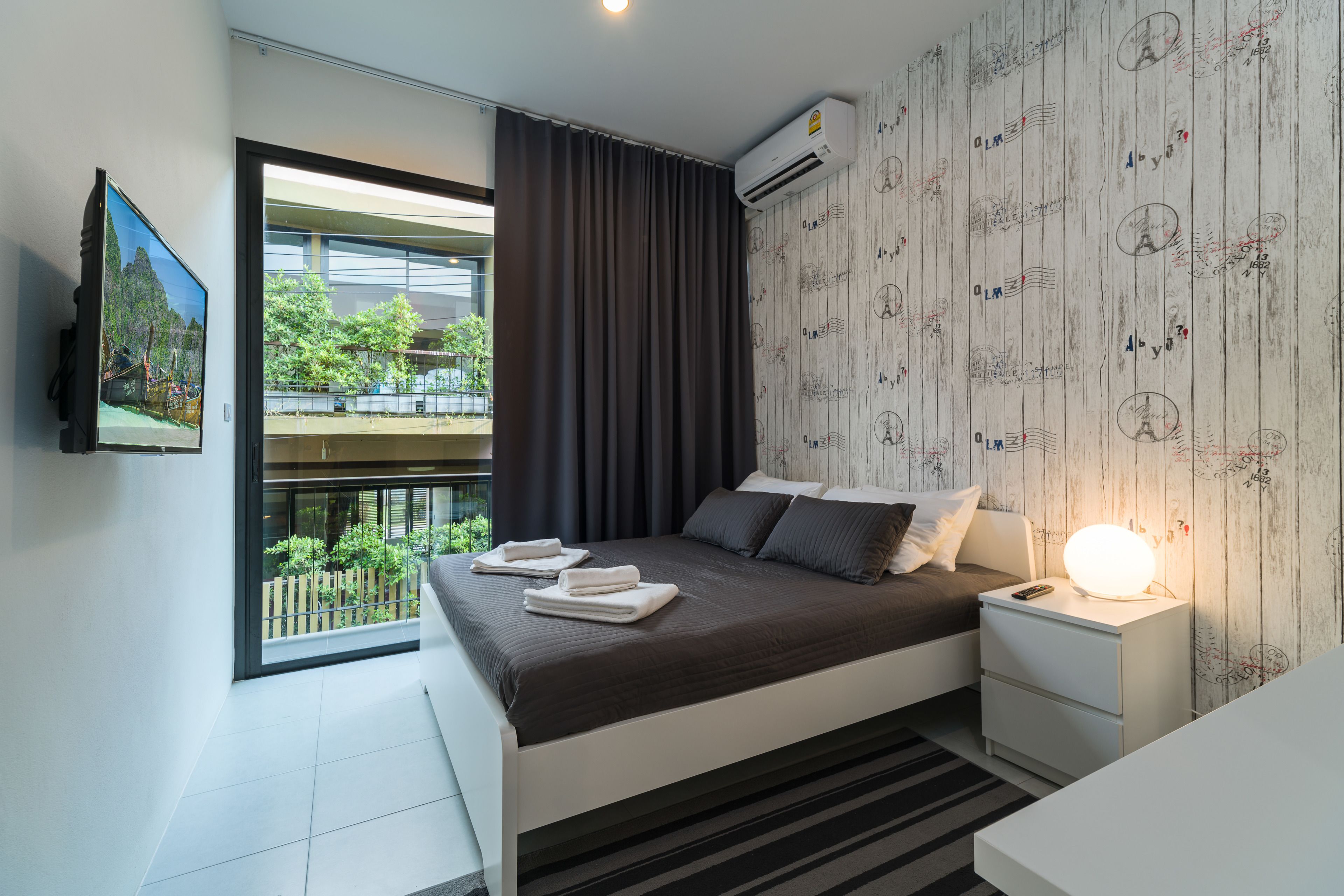 A modern bedroom with a large bed, a bedside table with a lamp, a TV on the wall, and a balcony with a view of greenery.