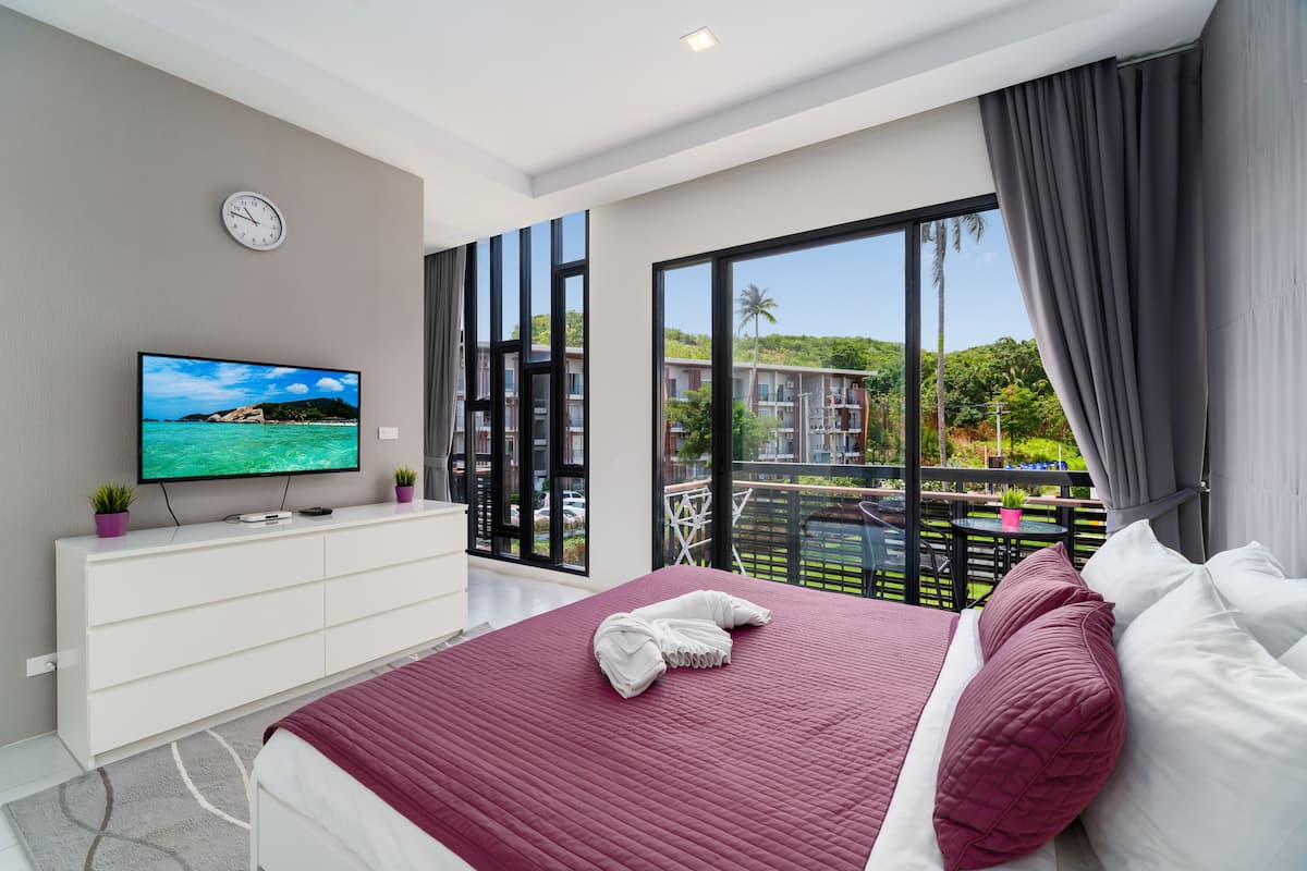 A modern bedroom with a large bed, a TV on a white dresser, and a balcony with a view of a resort.
