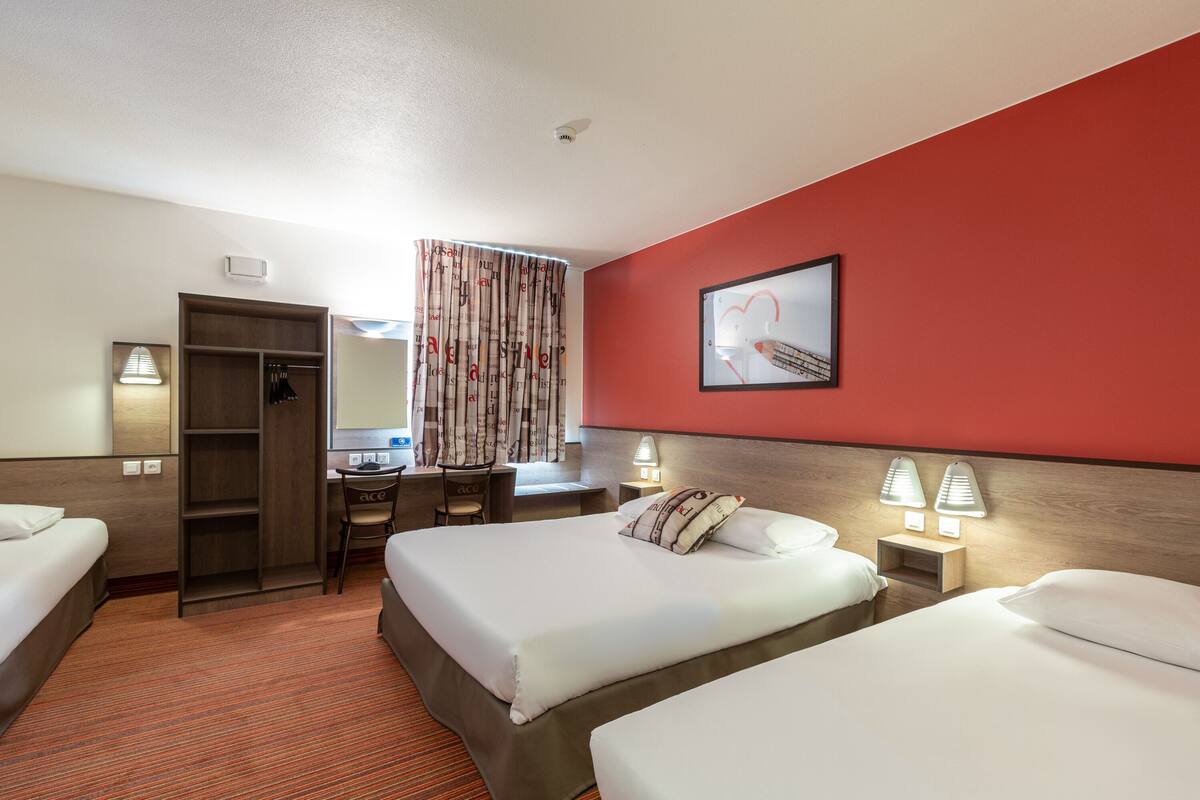 Une chambre d’hôtel avec deux lits, un bureau, une chaise, une armoire et un mur d’accent rouge.