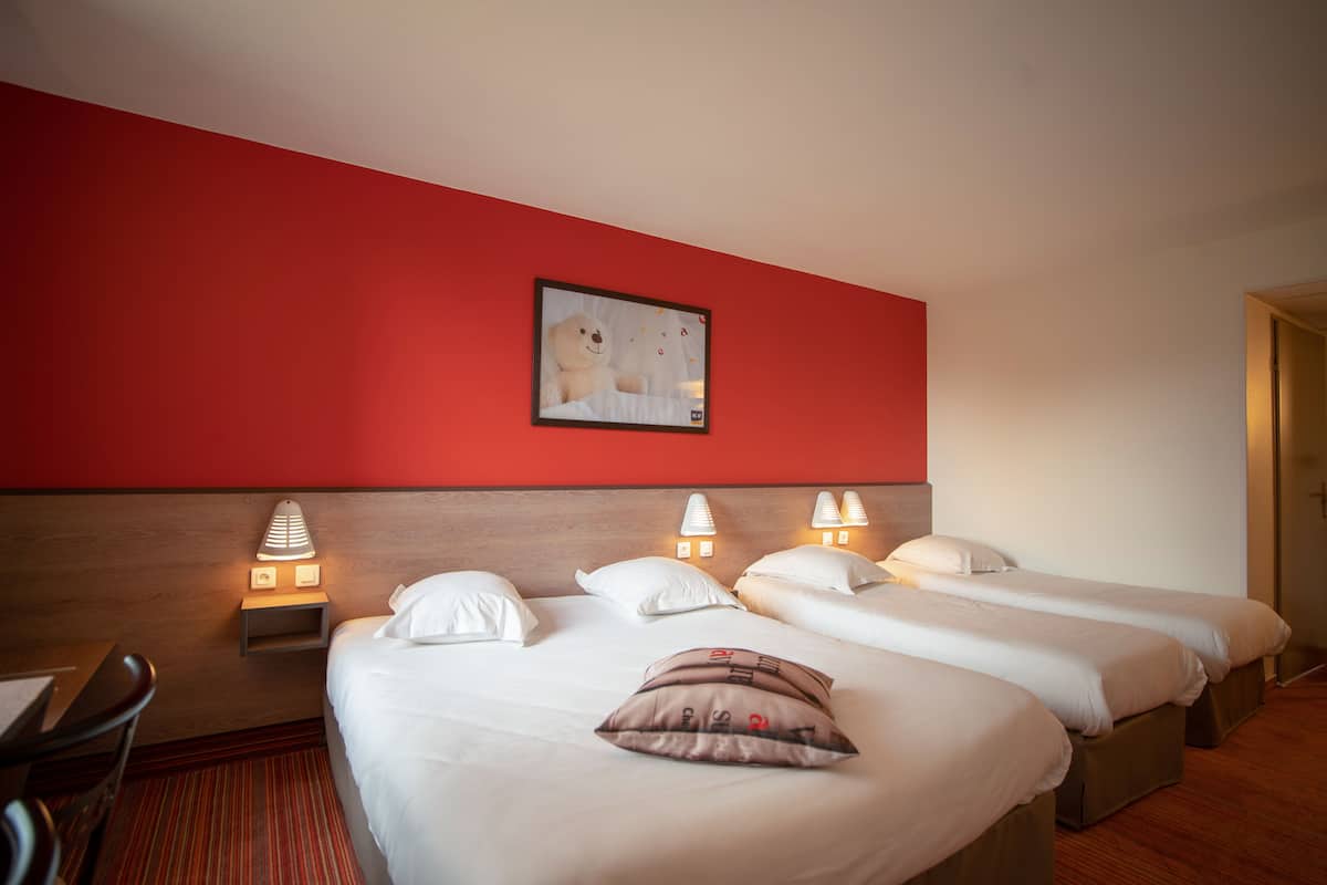 Une chambre d’hôtel avec trois lits simples, une tête de lit en bois, un mur d’accent rouge et des lampes de chevet.