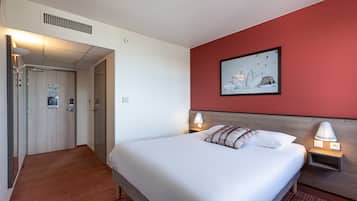 Habitación de hotel con cama, lámparas de noche, una pared de acento roja, un cuadro enmarcado y una puerta que da a otra habitación.
