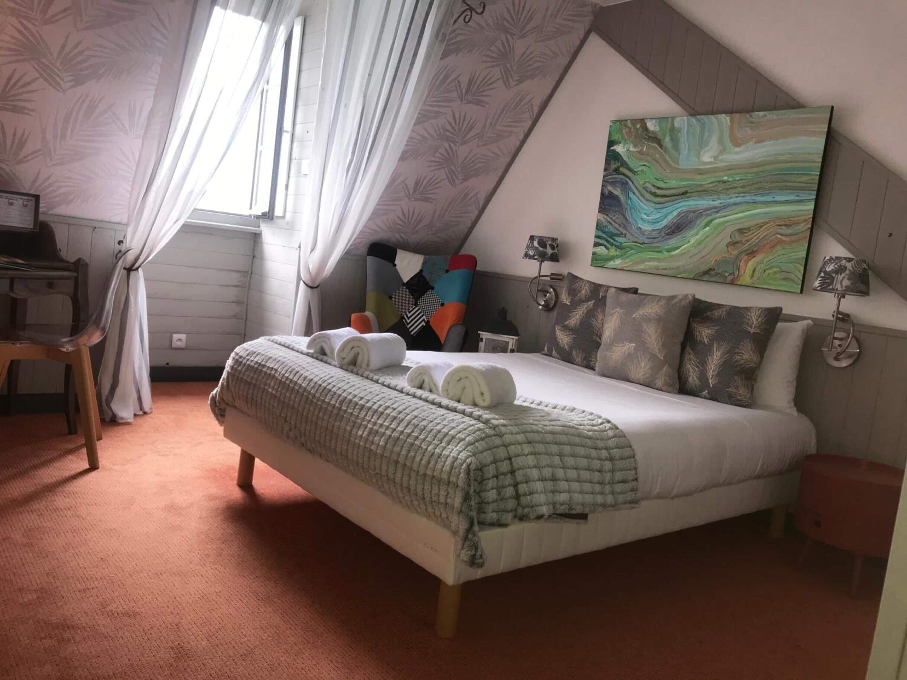 Une chambre avec un grand lit, un bureau et une chaise. Il y a une fenêtre avec des rideaux, un tableau au mur et une lampe sur la table de chevet.