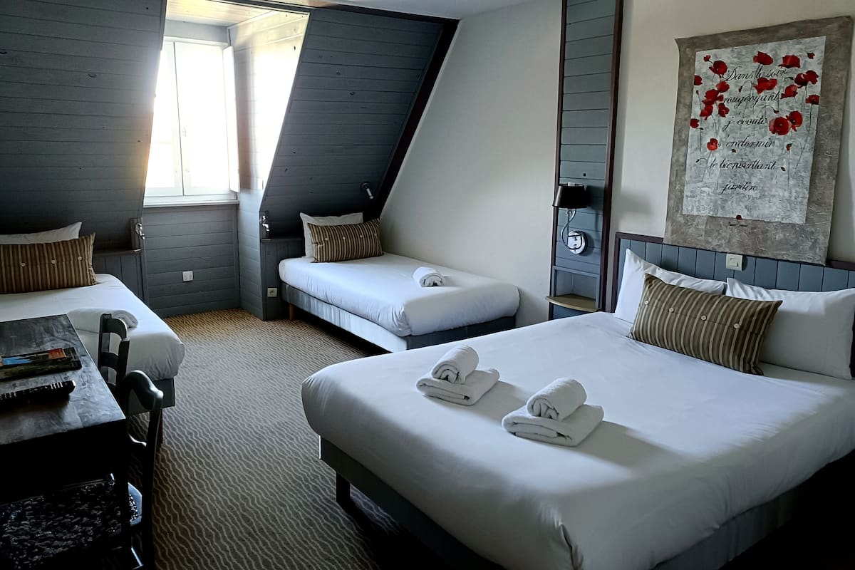 Une chambre d’hôtel avec deux lits simples, un grand lit, un bureau avec une chaise, une fenêtre et une lampe fixée au mur.