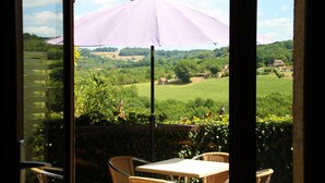 A patio with a table and chairs, a purple umbrella, and a view of a green landscape through a window. - Hôtel Bien-Etre Aux Cyprès de Marquay (Marquay)