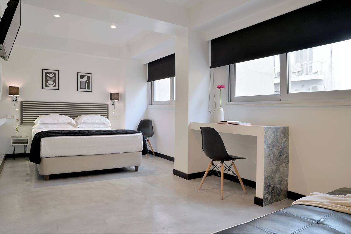 A modern hotel room with a large bed, a desk, and a chair. There are two framed pictures on the wall, a window with a view of buildings, and a vase with flowers on the desk.