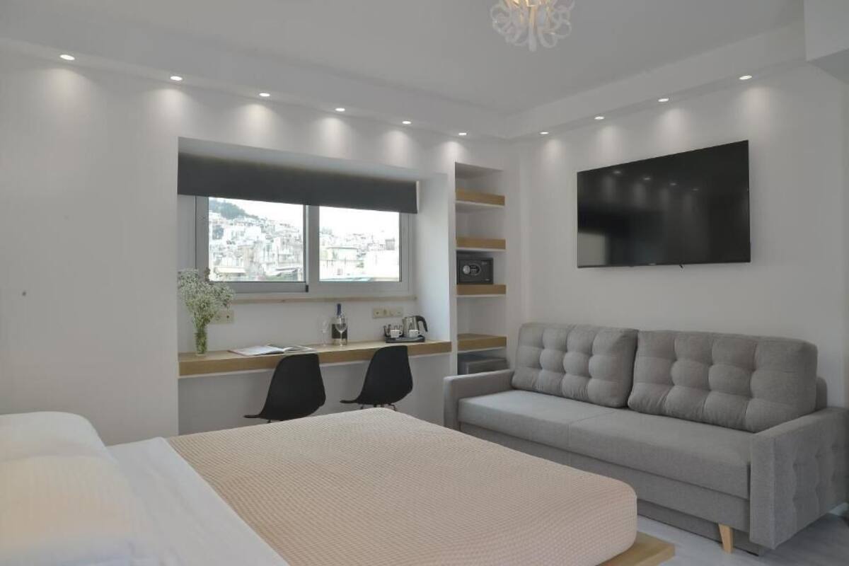A modern hotel room with a large bed, a sofa, a desk with a chair, a TV, and a window with a city view.